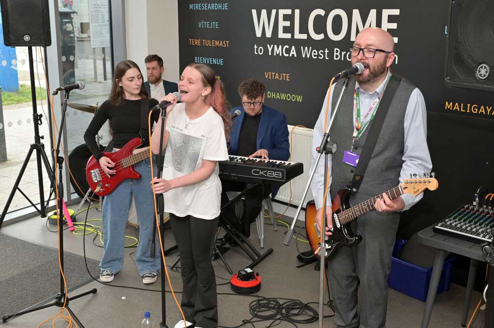 A mixed group of teachers and students from a local school, each with different instruments performing in YMCA cafe. The lead singer is a young, white, female.